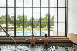 Deux personnes dans une piscine, avec vue sur un espace aménagé au bord de la piscine et des parasols.