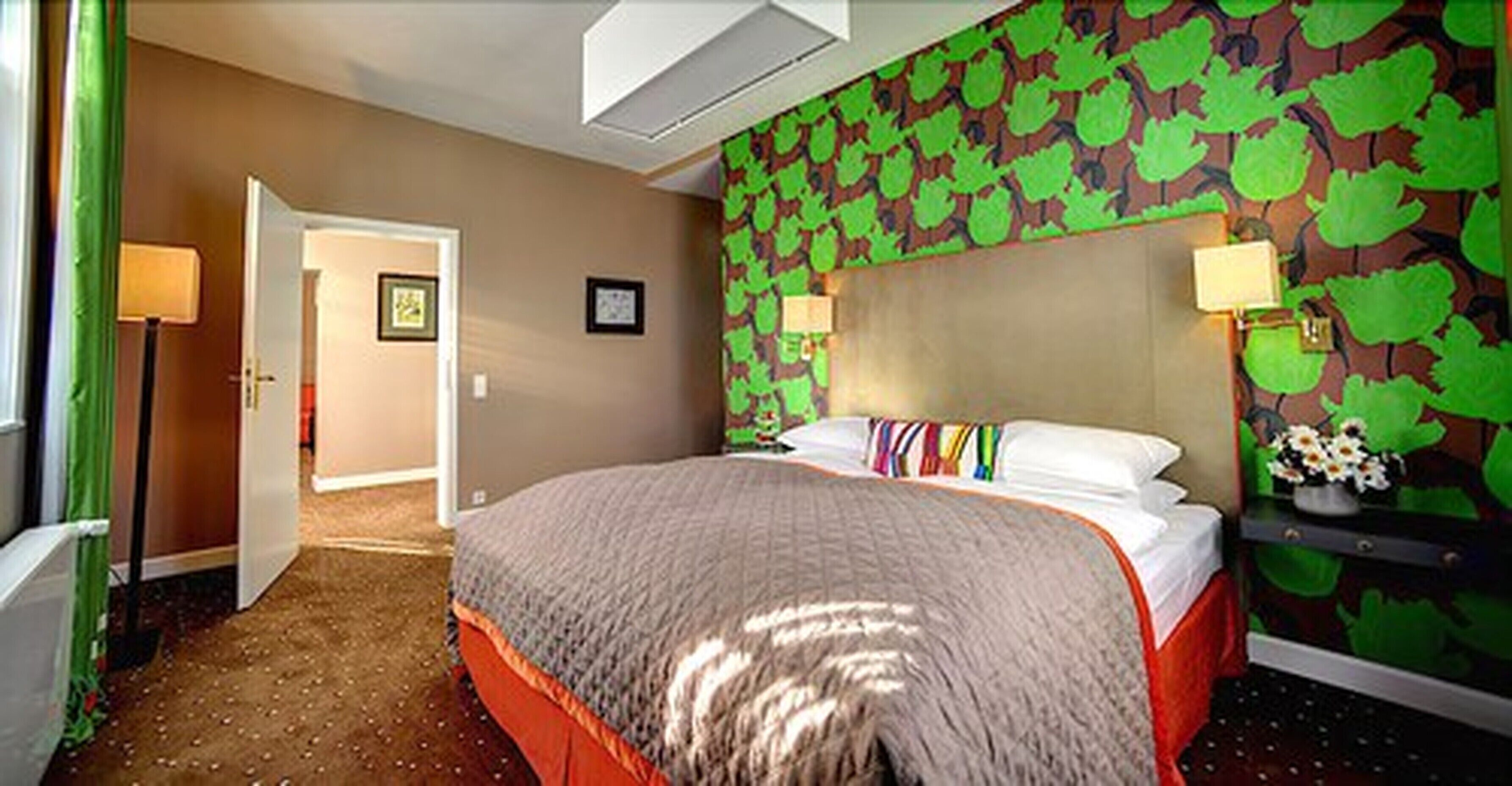A hotel room with a bed, a headboard with a leaf pattern, a nightstand with a lamp, and a door leading to another room.