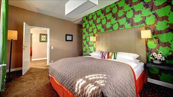 A hotel room with a bed, a headboard with a leaf pattern, a nightstand with a lamp, and a door leading to another room.