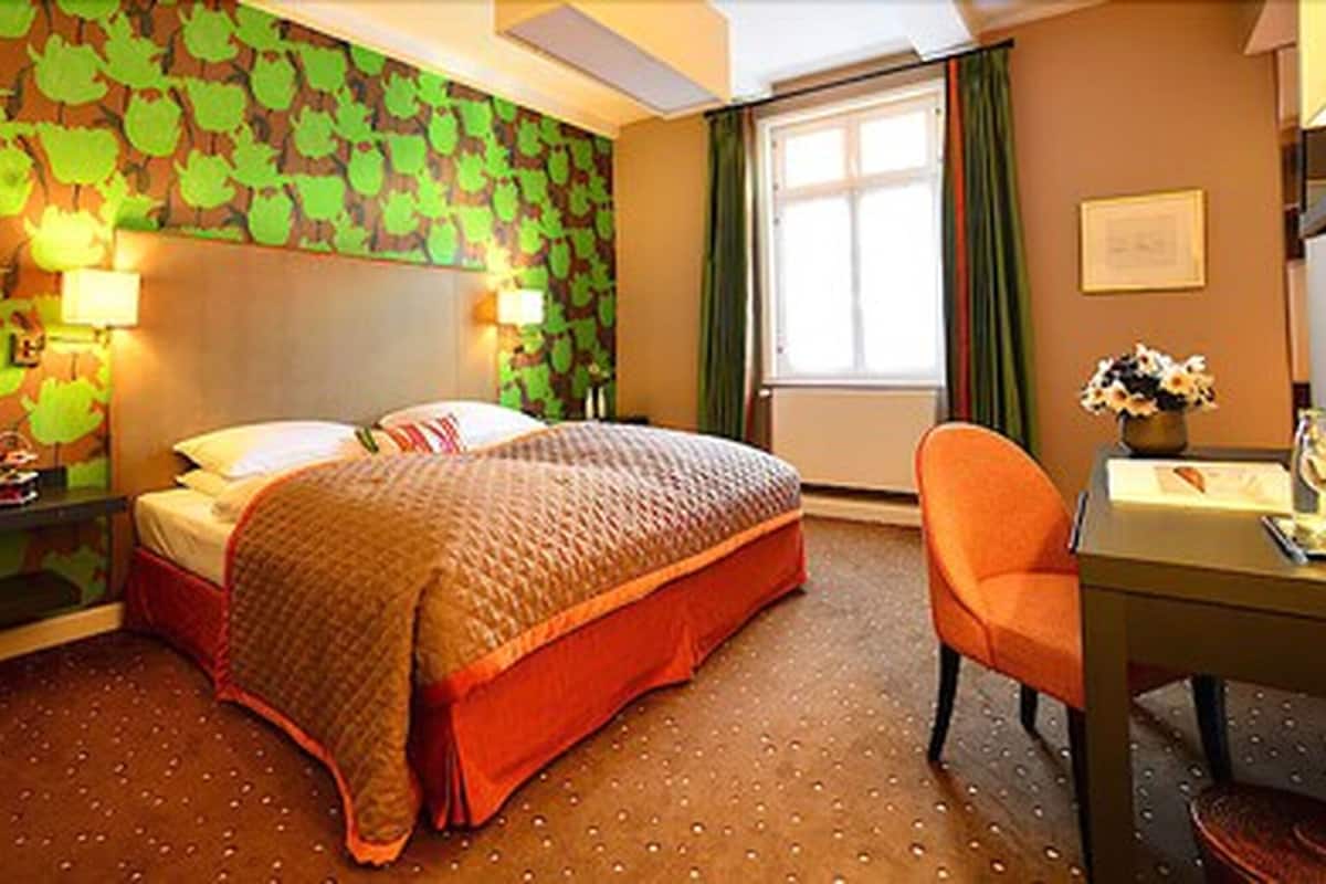 Ein Hotelzimmer mit einem Bett, einem Schreibtisch, einem Stuhl, einem Fenster und einer Wand mit einem Blattmuster.