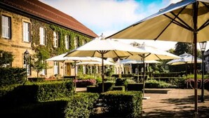 Een terras buiten met parasols, een gebouw met een rood dak en keurig onderhouden hagen.
