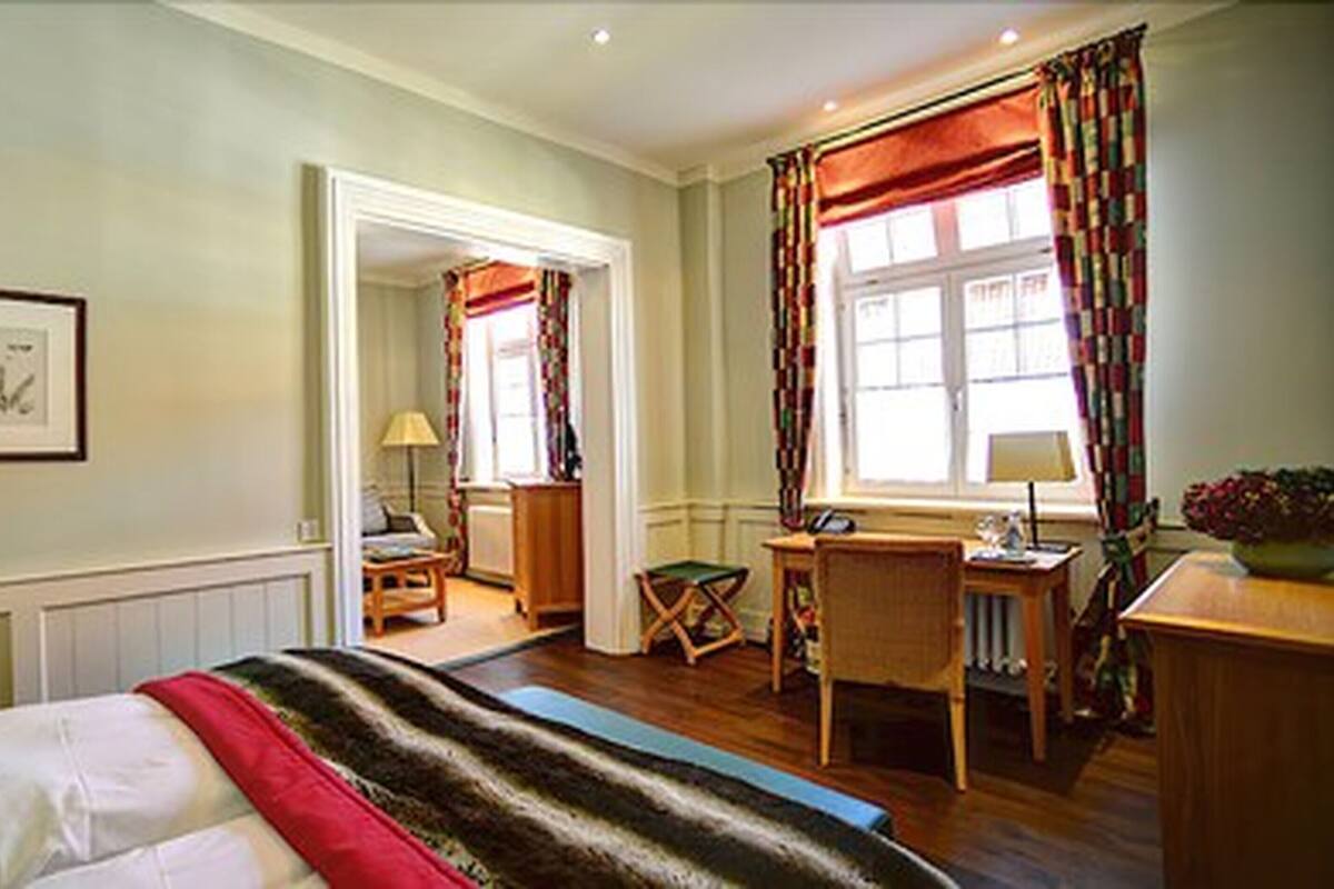 Ein Schlafzimmer mit einem Bett, einem Schreibtisch und einem Stuhl. Ein Fenster mit Vorhängen und ein Bild an der Wand sind zu sehen.