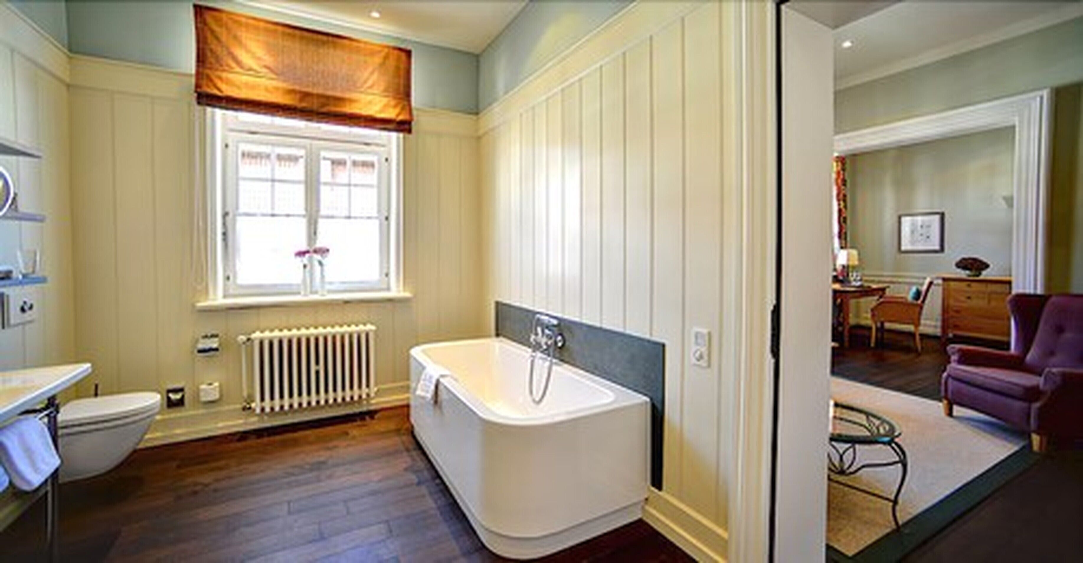 Une salle de bain avec une baignoire, des toilettes, une fenêtre avec un rideau et un radiateur.