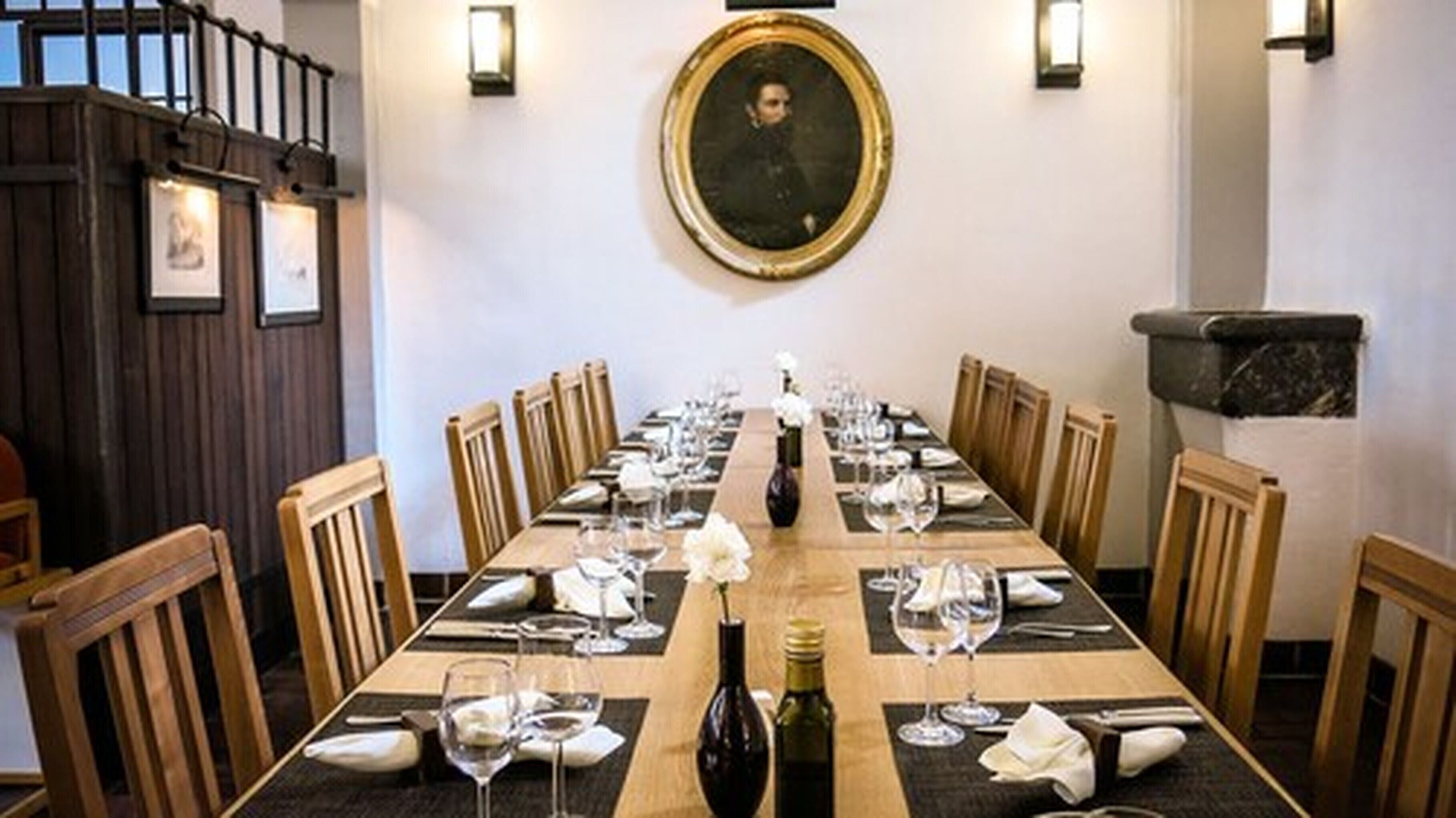 Une longue table à manger, dressée pour un repas, avec des chaises de chaque côté, un portrait encadré au-dessus de la table et un escalier en bois sur la gauche.