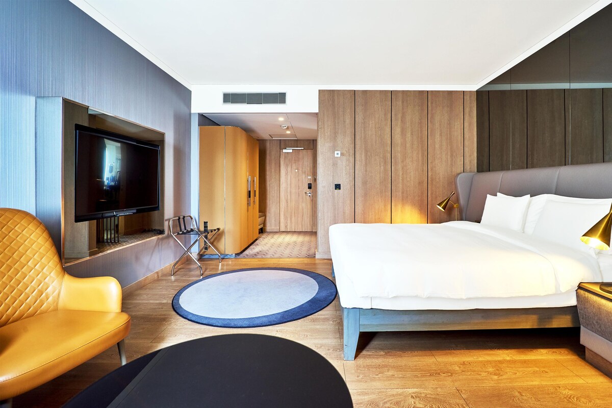 A modern hotel room with a large bed, a flat-screen TV, a leather armchair, and a wooden wardrobe.