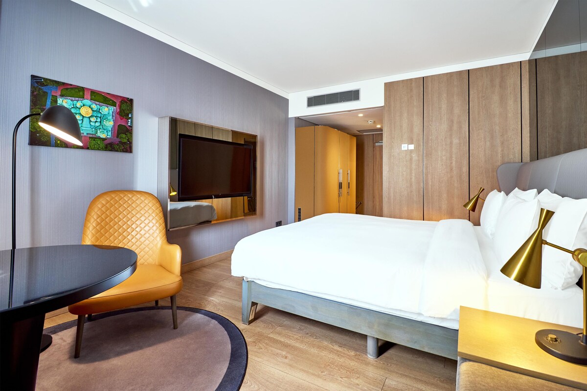 A modern hotel room with a bed, a desk, a chair, a TV mounted on the wall, and a painting.