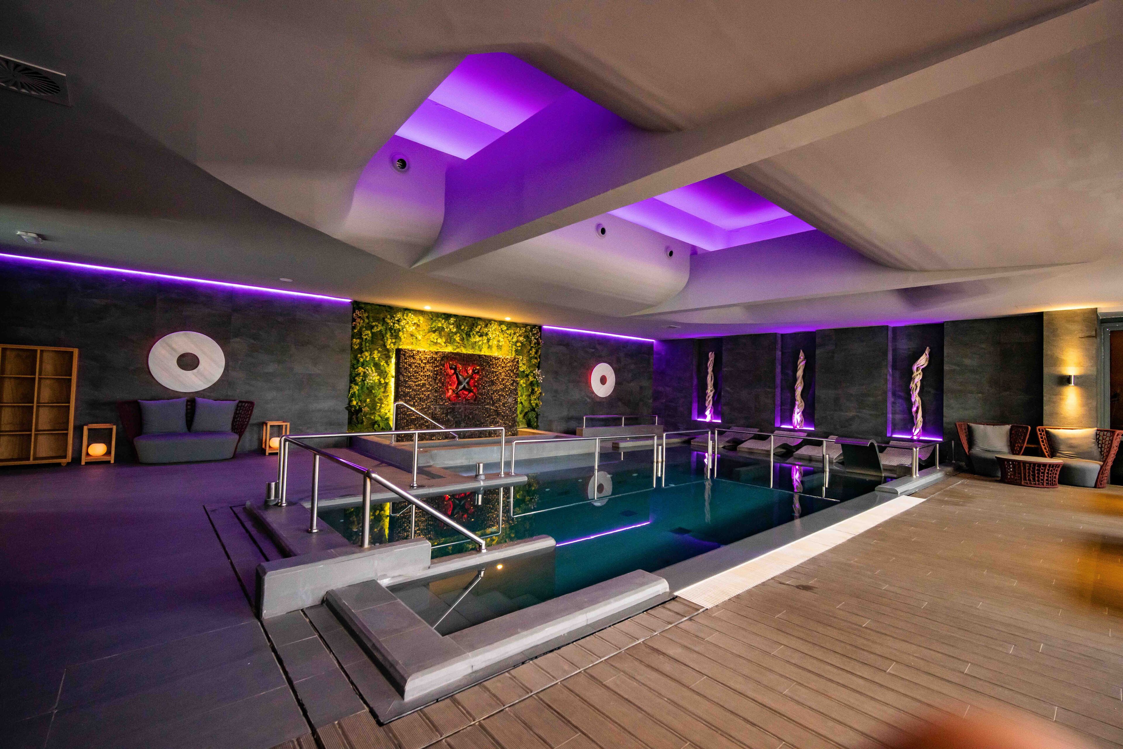 A modern indoor pool area with a wooden deck, a seating area with cushions, and a wall with a decorative plant and wall art.