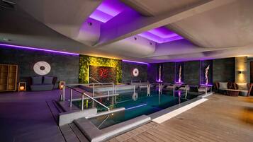Un área de piscina interior moderna con cubierta de madera, zona de estar con cojines y pared decorada con plantas y arte.