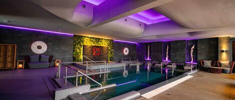 A modern indoor pool area with a wooden deck, a seating area with cushions, and a wall with a decorative plant and wall art.