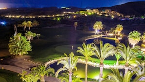 Vista nocturna de un campo de golf con palmeras, un estanque y edificios iluminados al fondo.