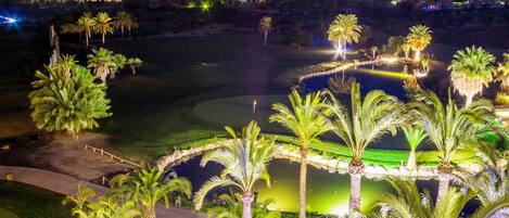 Vista nocturna de un campo de golf con palmeras, un estanque y edificios iluminados al fondo.