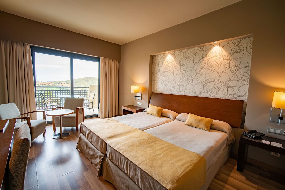 A hotel room with a large bed, a desk, a chair, a small table, and a view of the city.