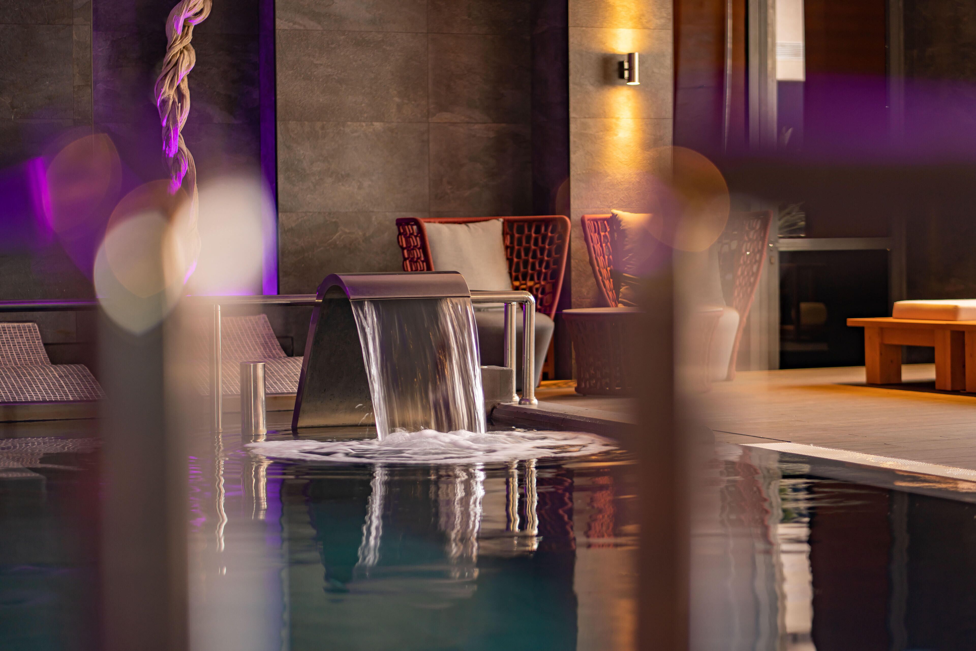 A modern indoor pool area with a waterfall feature, lounge chairs, and a seating area with cushions.