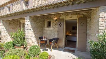 A stone building with a patio area featuring two wicker chairs and a small table, a door leading to a bedroom, and potted plants around the space.
