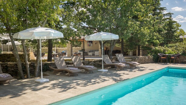 Zona de piscina con sillones de descanso y sombrillas, un muro de piedra y una alberca.