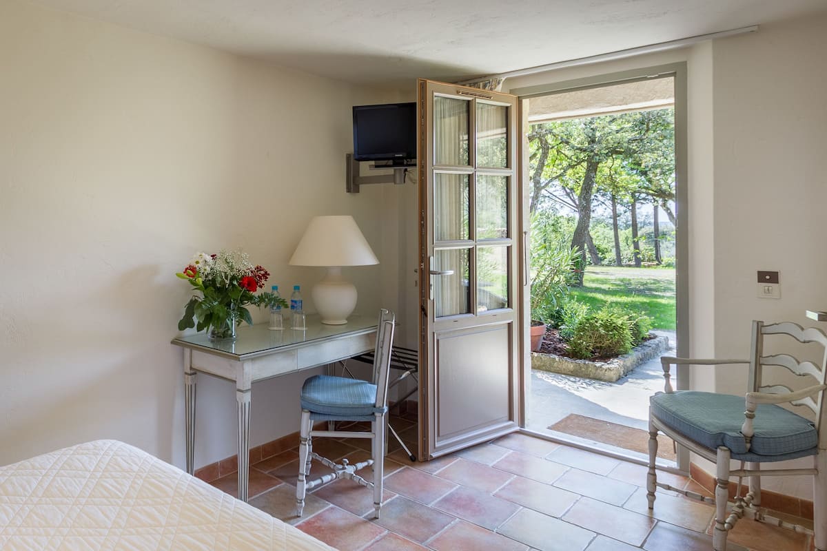 A room with a bed, a desk with a chair, a TV, and a door leading to an outdoor area with plants.