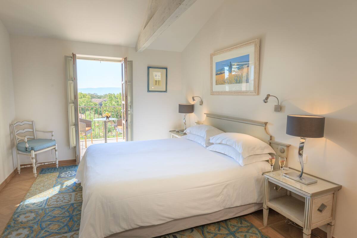 A bedroom with a large bed, two bedside tables with lamps, a chair, a balcony with a view, and a framed picture on the wall.