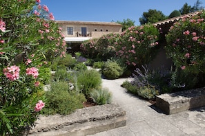 Un edificio de piedra con techo de tejas, rodeado por un jardín con plantas de flores rosadas y un sendero pavimentado.