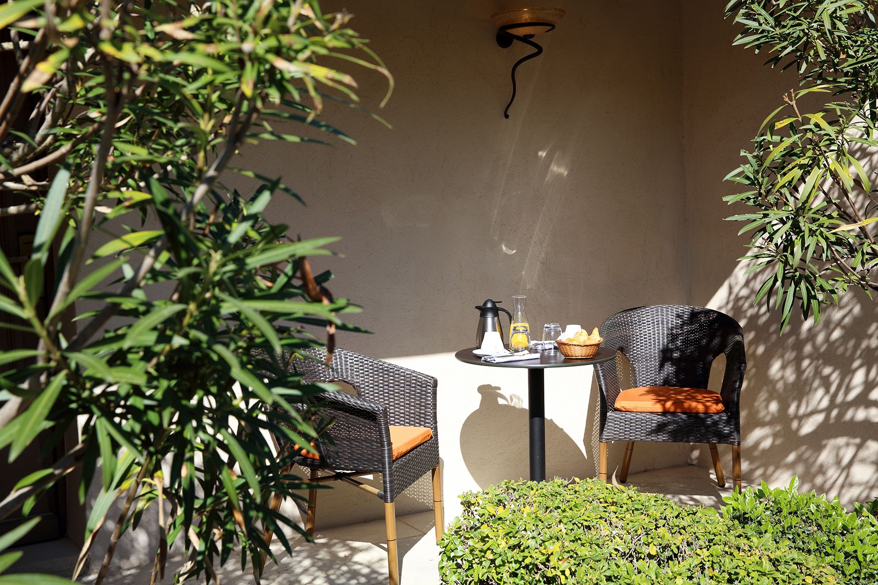 Uma pequena área externa com assento para dois, incluindo duas cadeiras de vime e uma mesa, uma cafeteira, um jarro de vidro e uma cesta de pão sobre a mesa.