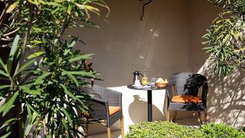 A small outdoor seating area with two wicker chairs and a table, a coffee pot, a glass pitcher, and a basket of bread on the table.