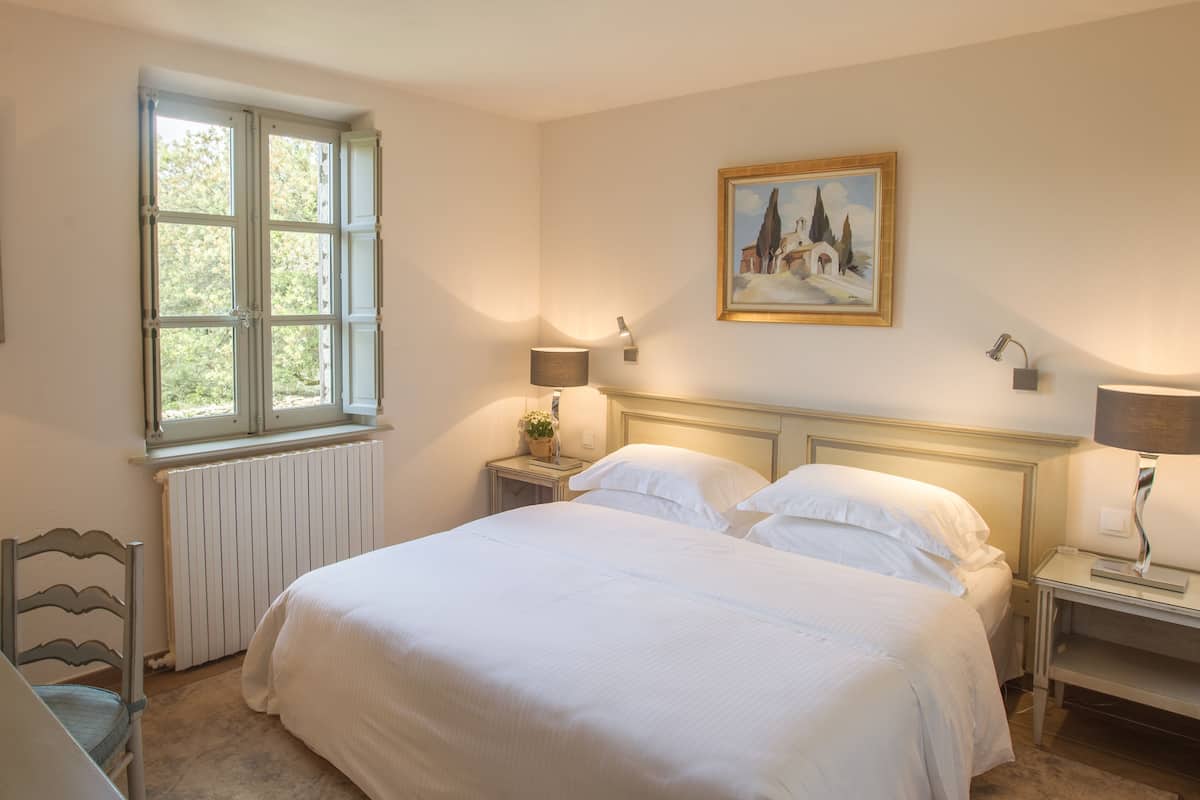 A bedroom with a large bed, a window with shutters, a radiator, bedside tables with lamps, and a painting on the wall.