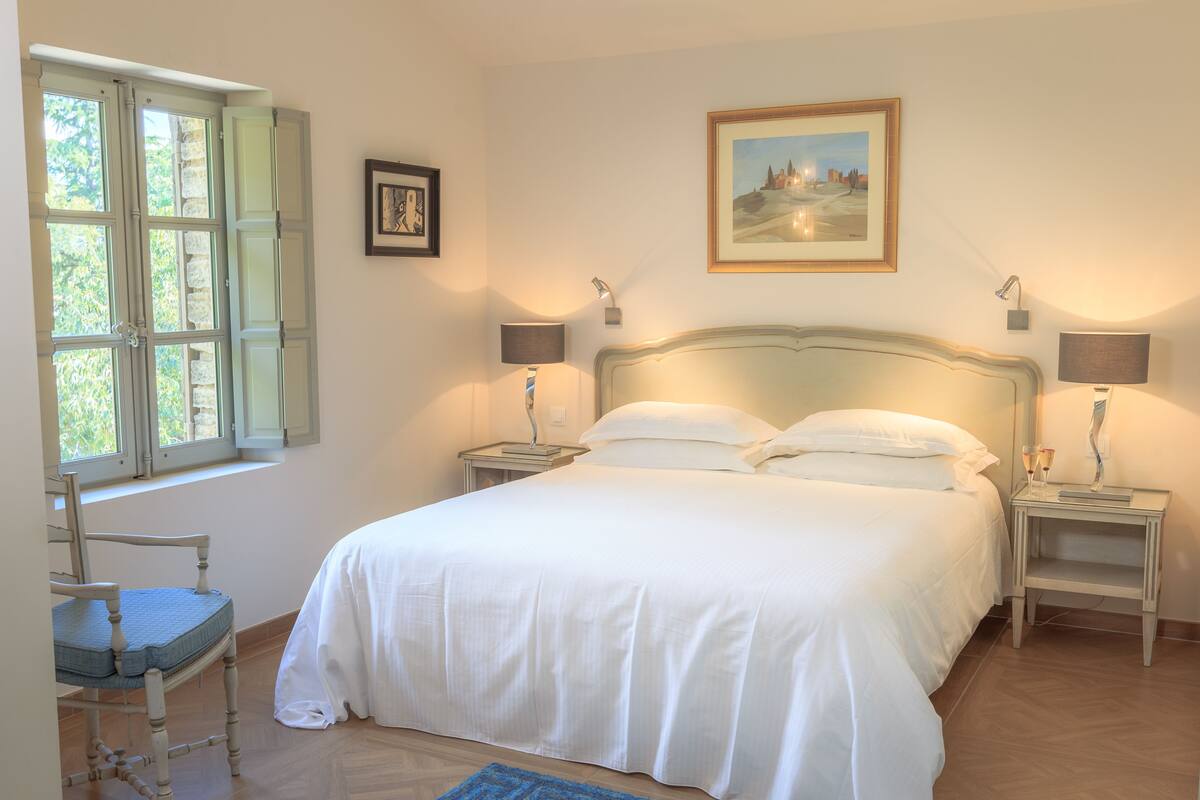 A bedroom with a large bed, two bedside tables with lamps, a chair, a window with shutters, and a framed picture on the wall.