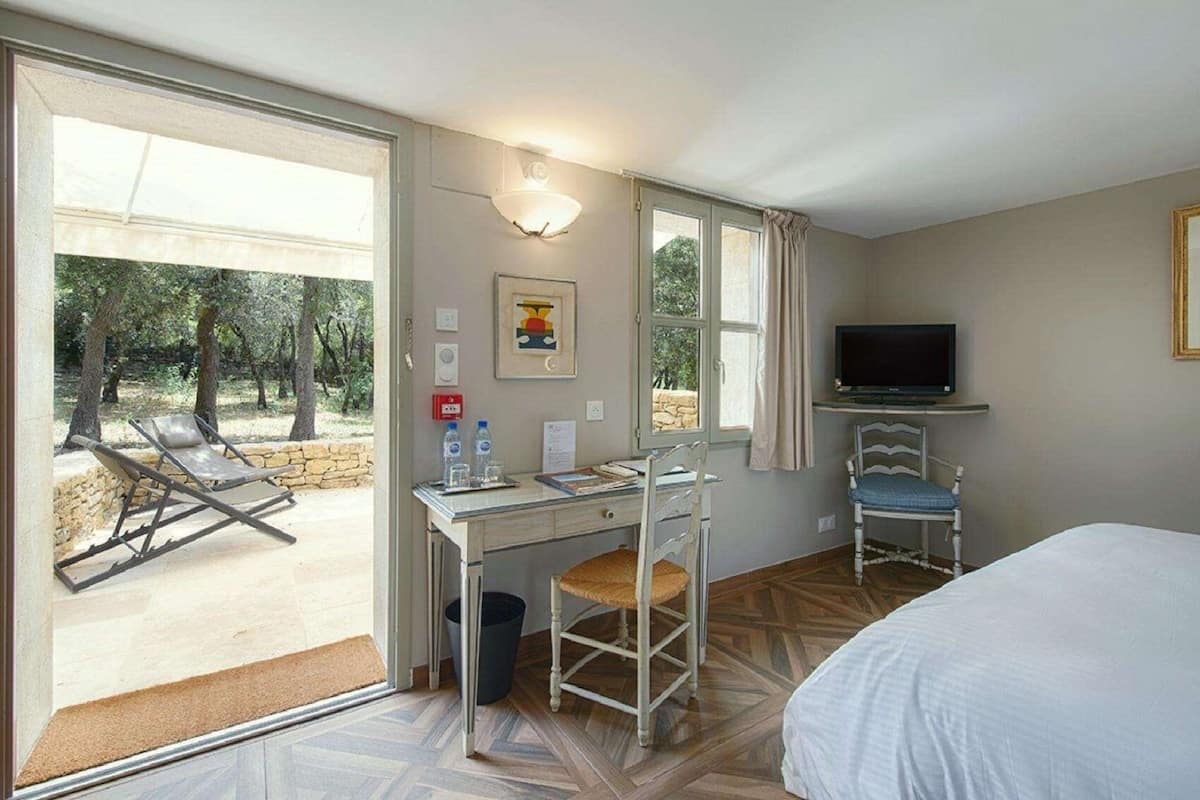 A hotel room with a bed, a desk with a chair, a television, and a view of an outdoor area with a sun lounger and trees.