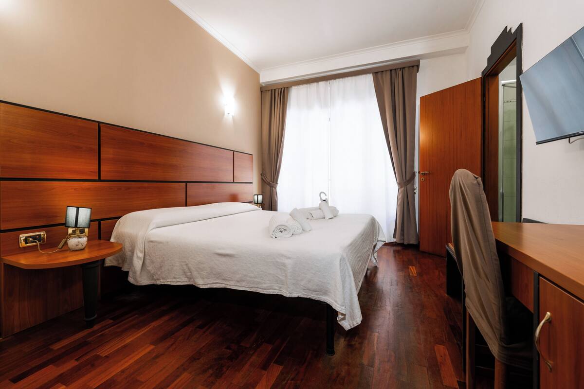 Una habitación de hotel con cabecera de madera, cama con ropa blanca, mesita de noche con lámpara, armario y televisor en la pared.