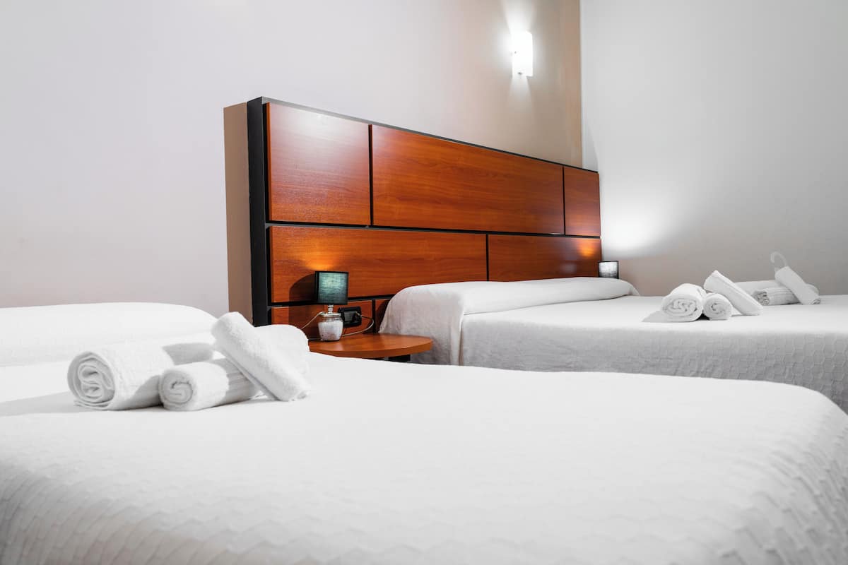 Habitación de hotel con dos camas, cabecero de madera, mesita de noche con lámpara y toallas enrolladas sobre las camas.