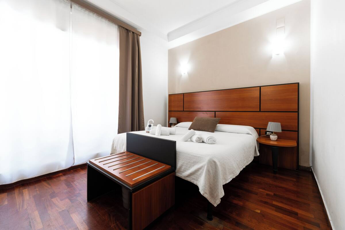 Habitación de hotel con cabecera de madera, cama con ropa blanca, mesita de noche con lámpara y un pequeño banco de madera.