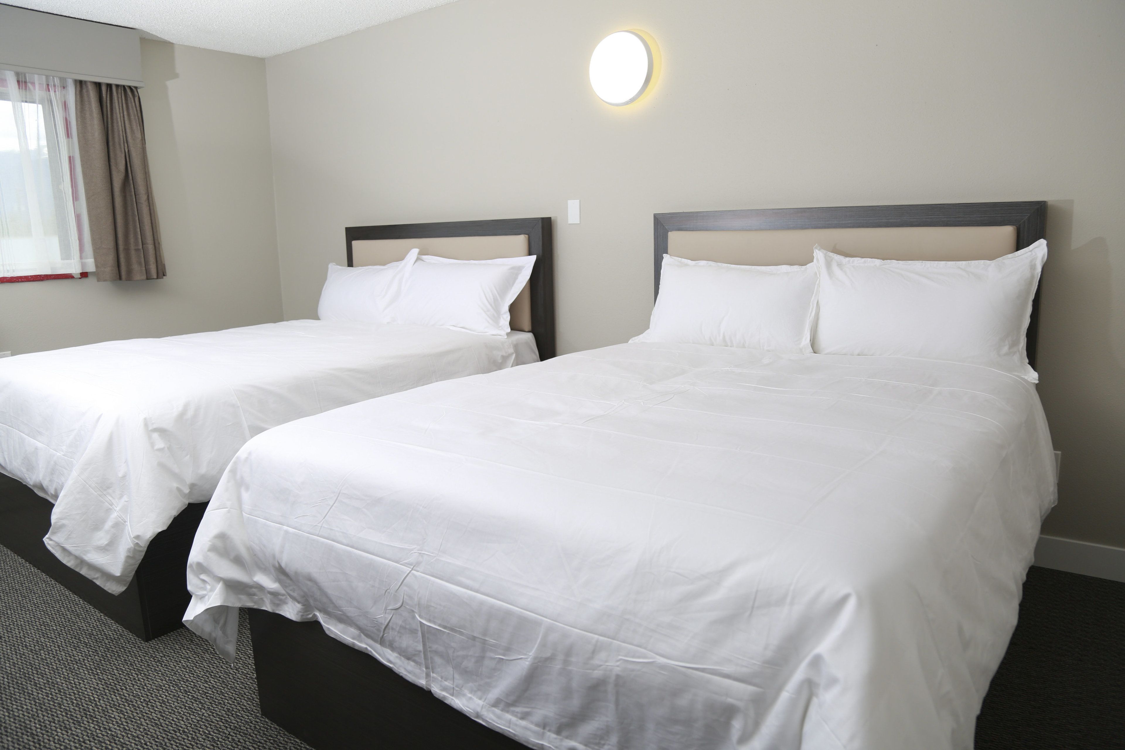 A hotel room with two single beds, a window with curtains, and a wall-mounted light fixture.