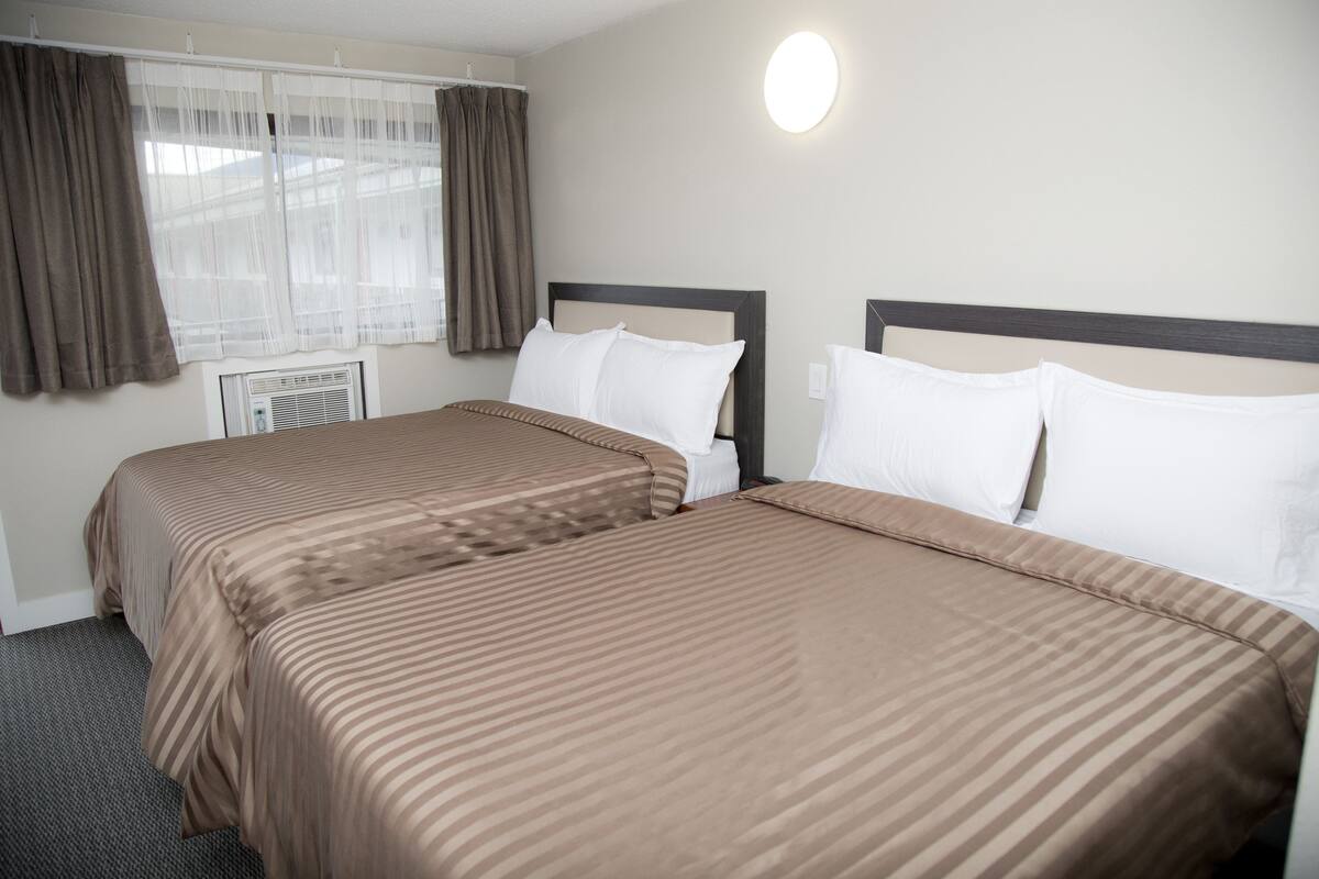 A hotel room with two beds, a window with curtains, a wall-mounted light, and a radiator.