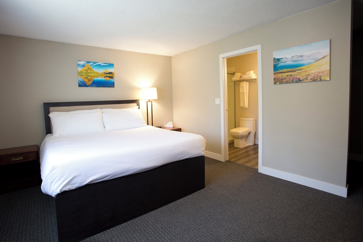A hotel room with a bed, a nightstand, a lamp, a painting, and a bathroom visible through an open door.