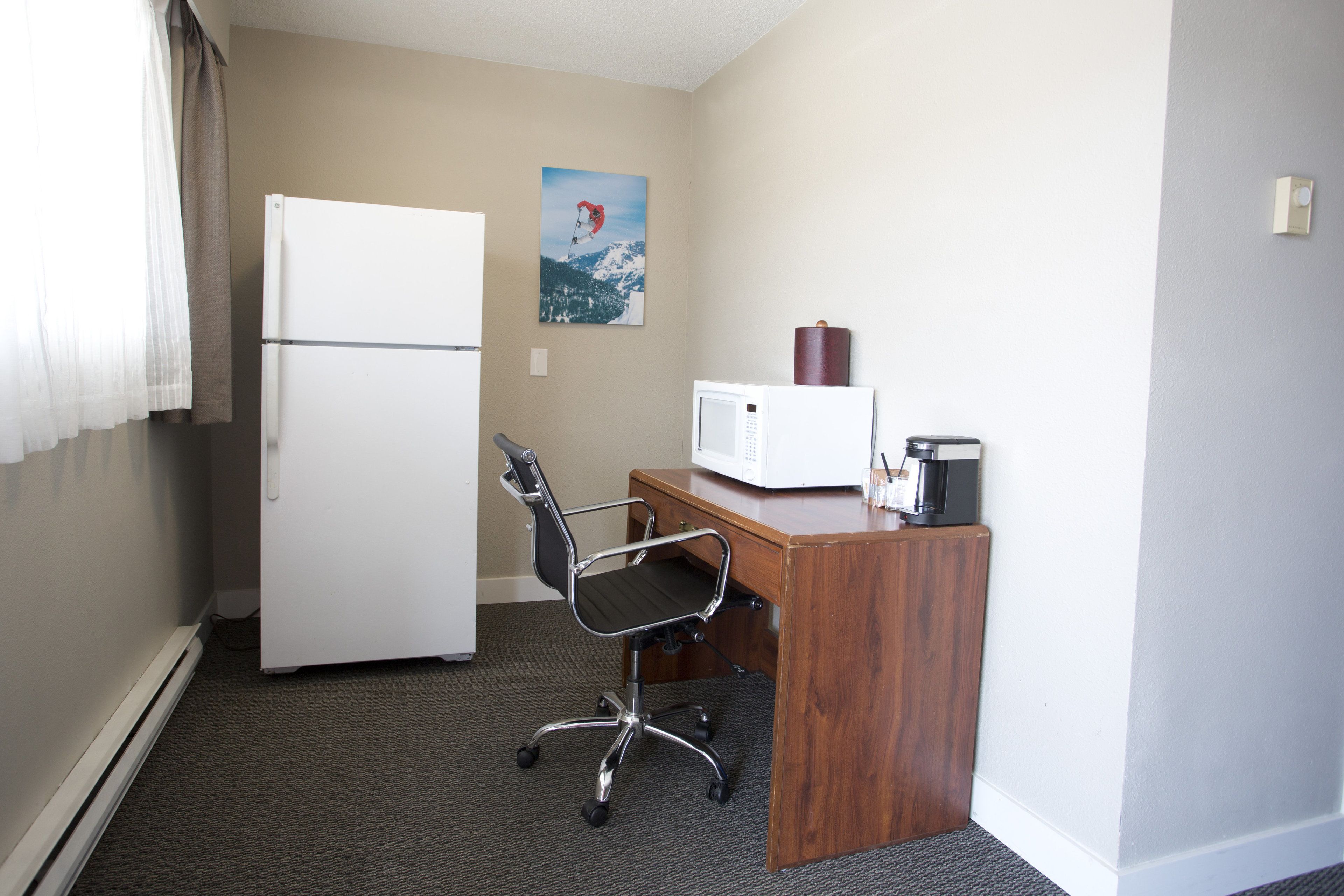 Une petite pièce propre, avec un réfrigérateur blanc, un bureau en bois équipé d’un ordinateur et d’une machine à café, une chaise de bureau noire et un tableau accroché au mur.