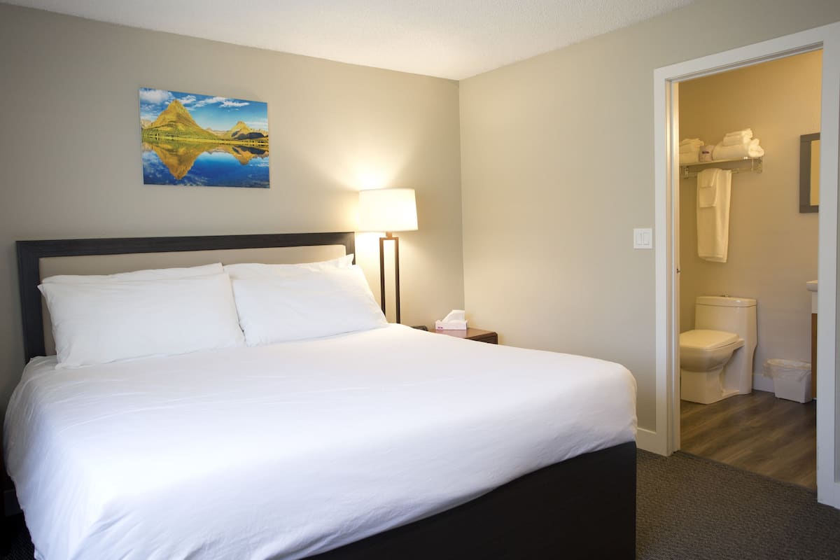 A hotel room with a bed, a nightstand, a lamp, a painting on the wall, and a bathroom visible through an open door.