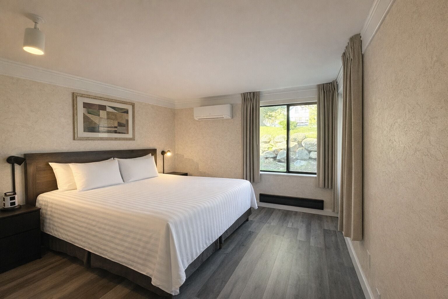 Ein Hotelzimmer mit einem großen Bett, einem Nachttisch, einer Wand-Klimaanlage, einem Fenster mit Vorhängen und einem gerahmten Bild an der Wand.