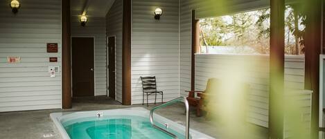 A covered outdoor swimming pool area with a hot tub, benches, and emergency exit signs.