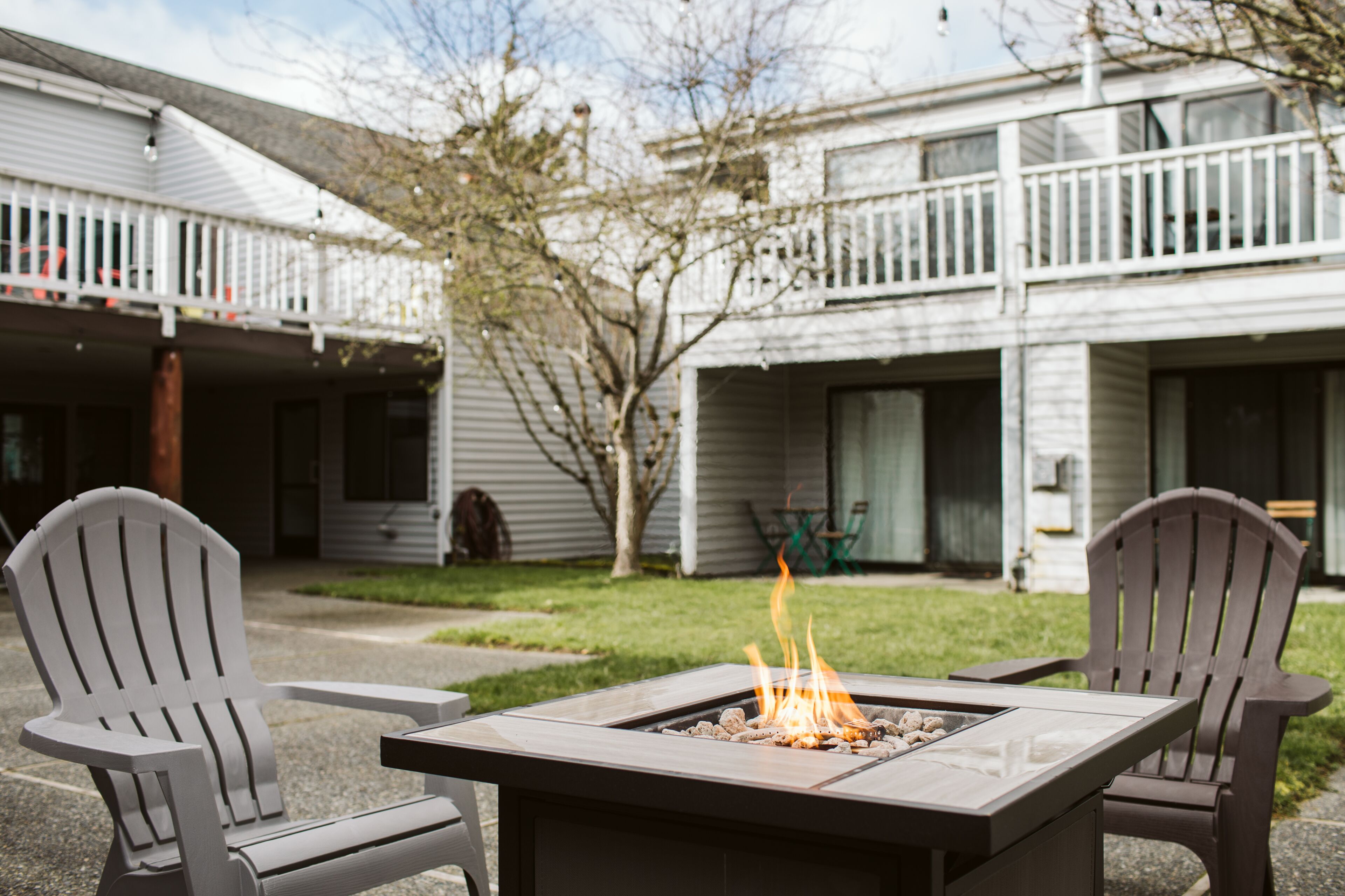 A patio with a fire pit and Adirondack chairs, a white building with a balcony, and a tree in the background.