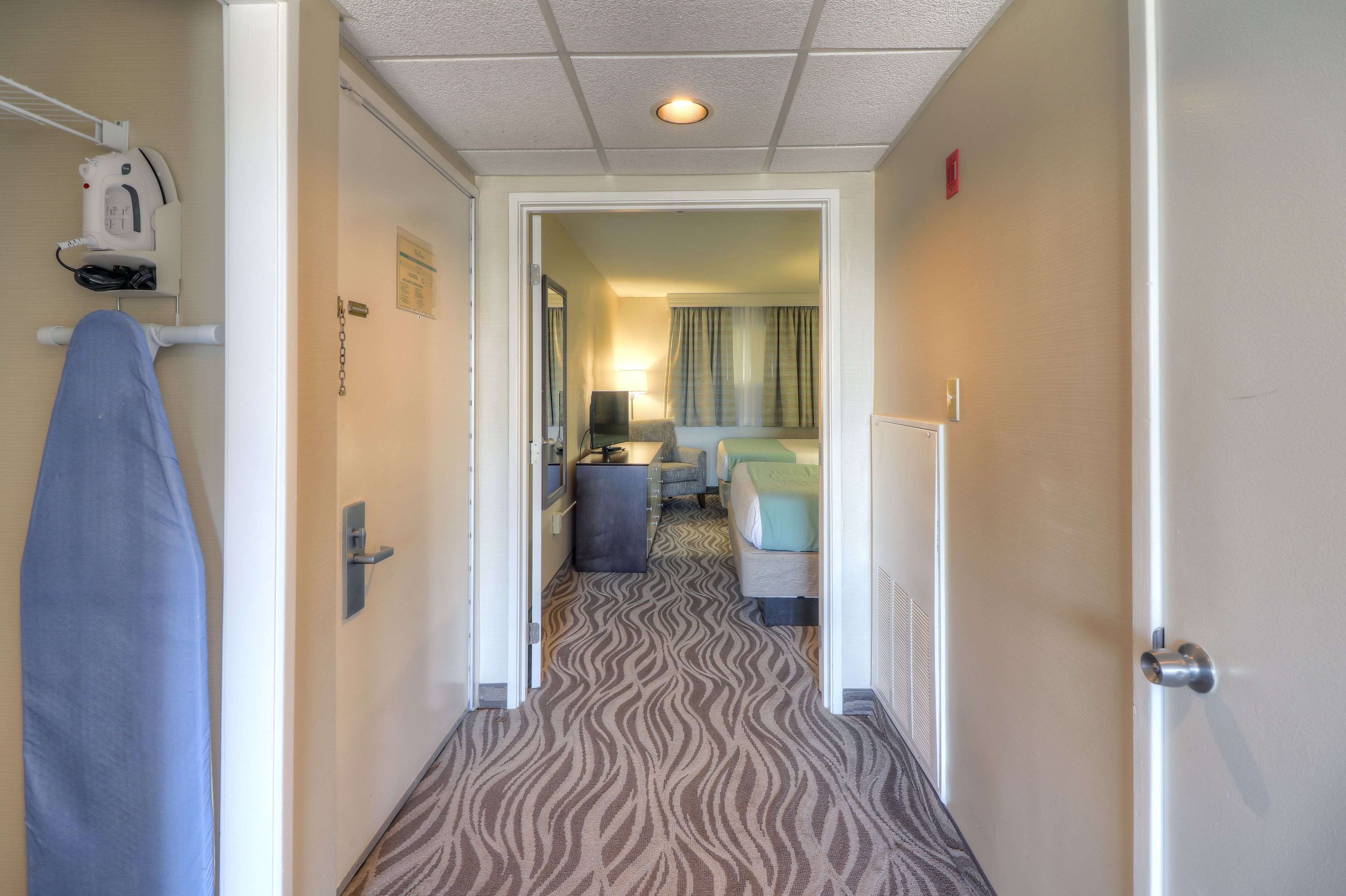 A hotel corridor with a patterned carpet, a bed, a desk, and a television.
