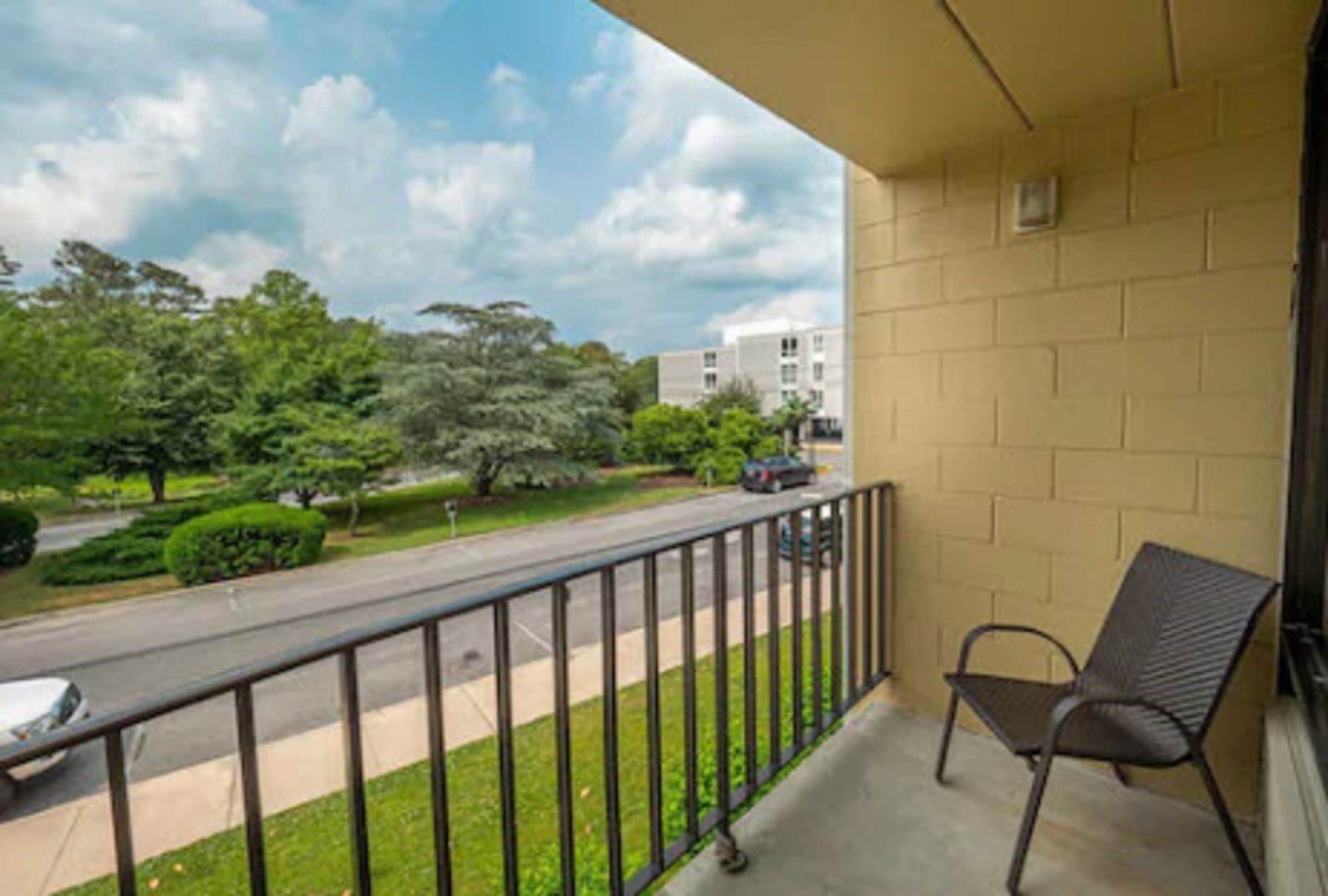 A balcony with a metal railing, a chair, and a view of a road and greenery.