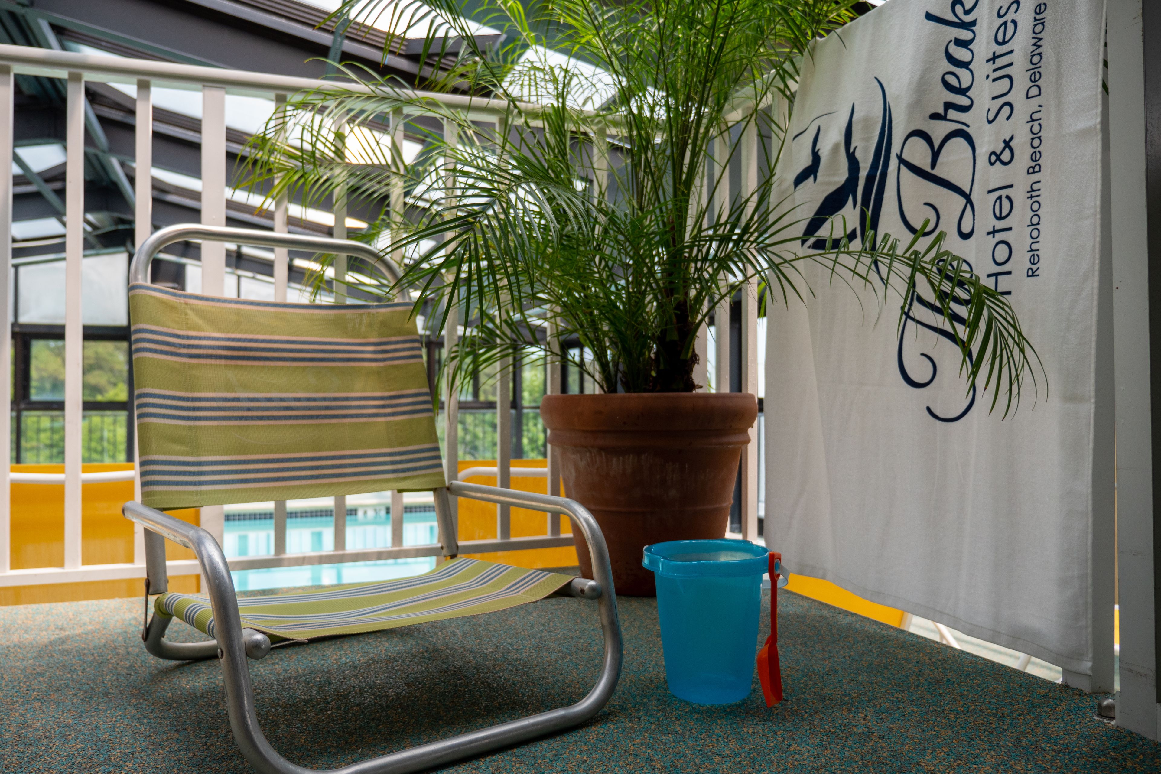 A chair with a striped cushion, a potted plant, and a blue bucket on a balcony with a railing and a sign for The Break Hotel & Suites.