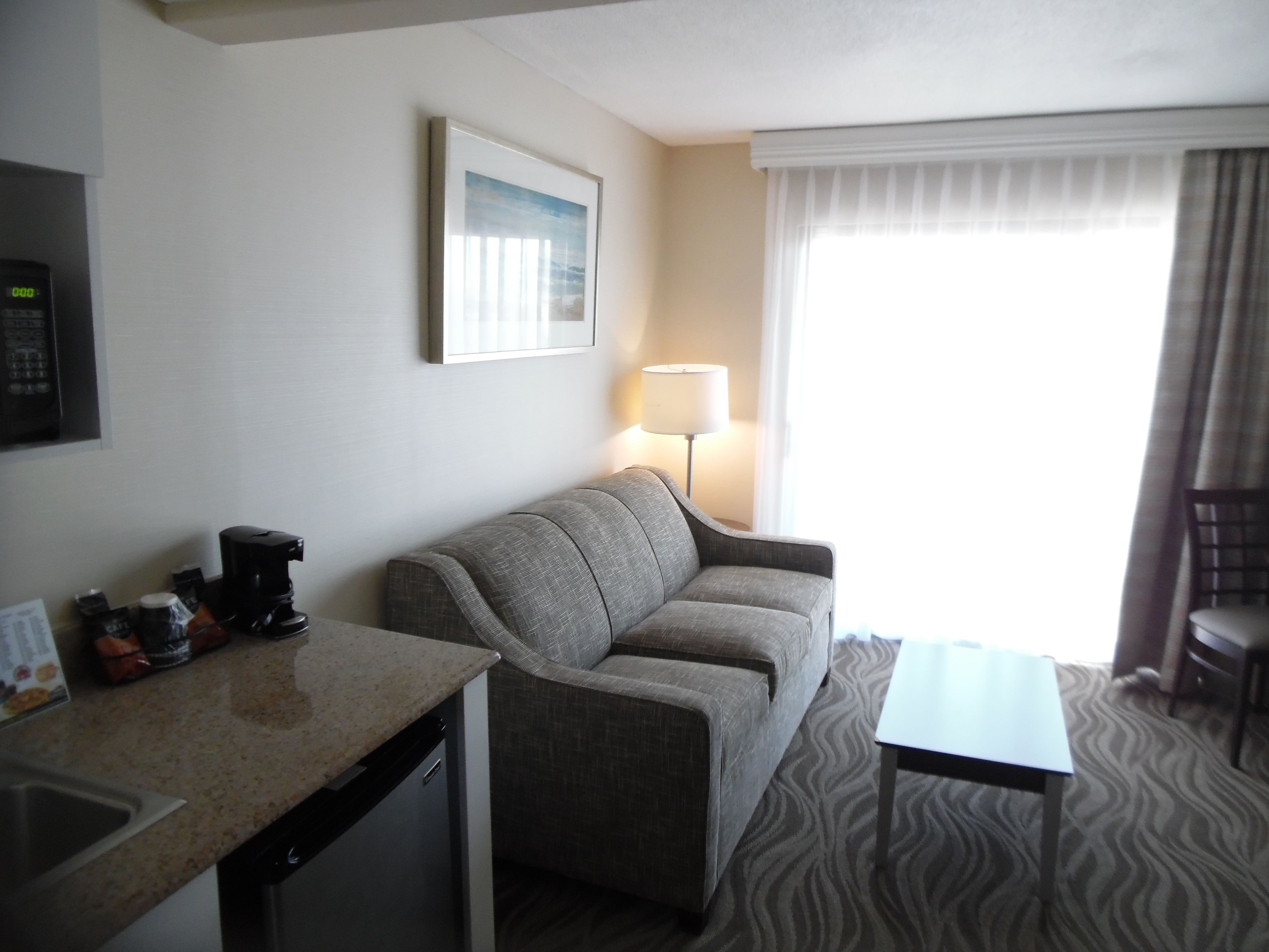 A hotel room with a kitchenette, a grey sofa, a small table, a lamp, and a window with curtains.