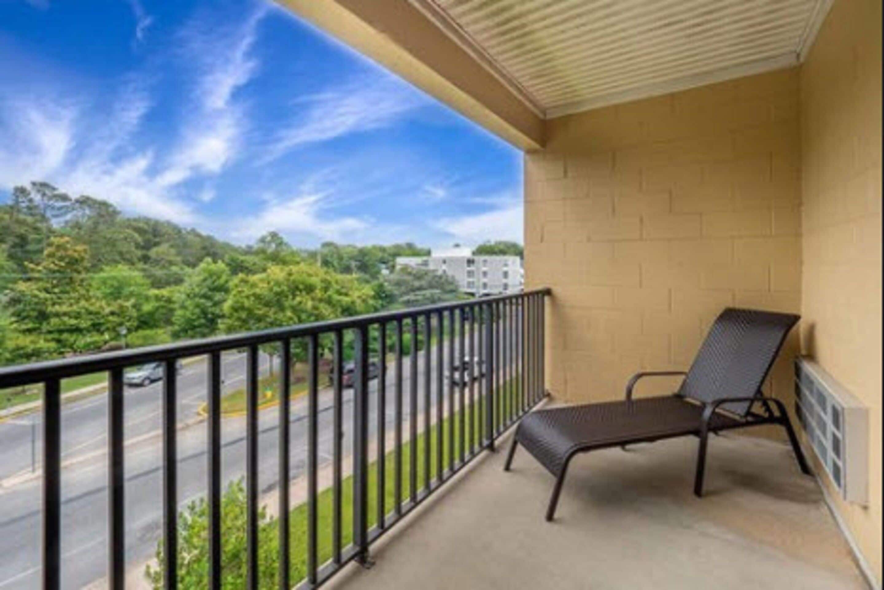 A balcony with a black metal chair, beige brick walls, and a view of a road and greenery.