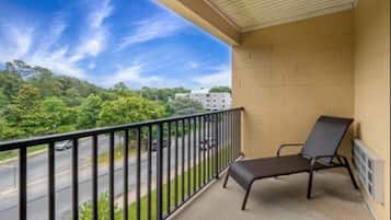 A balcony with a black metal chair, beige brick walls, and a view of a road and greenery.