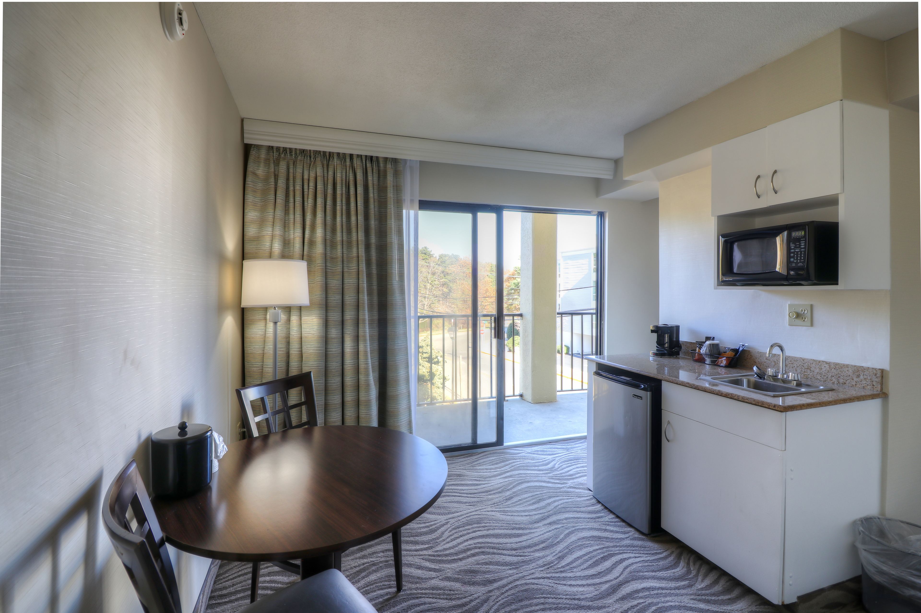 Ein kleines Hotelzimmer mit Esstisch, Stühlen, einer Mikrowelle, einem Spülbecken und Blick ins Freie durch eine Schiebetür.