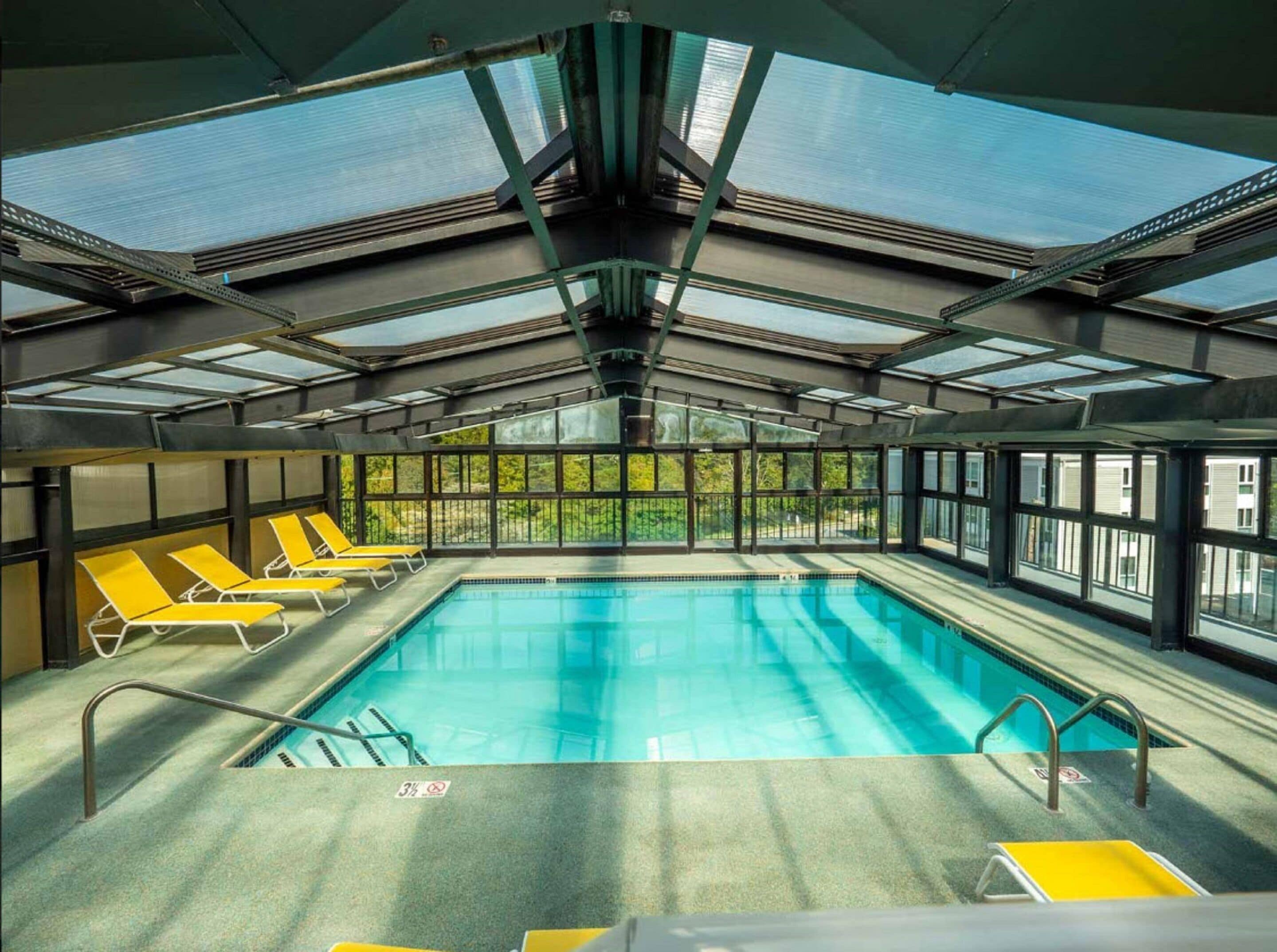 Une piscine intérieure dotée d’un toit en verre transparent, de chaises longues jaunes et offrant une vue sur la verdure environnante.