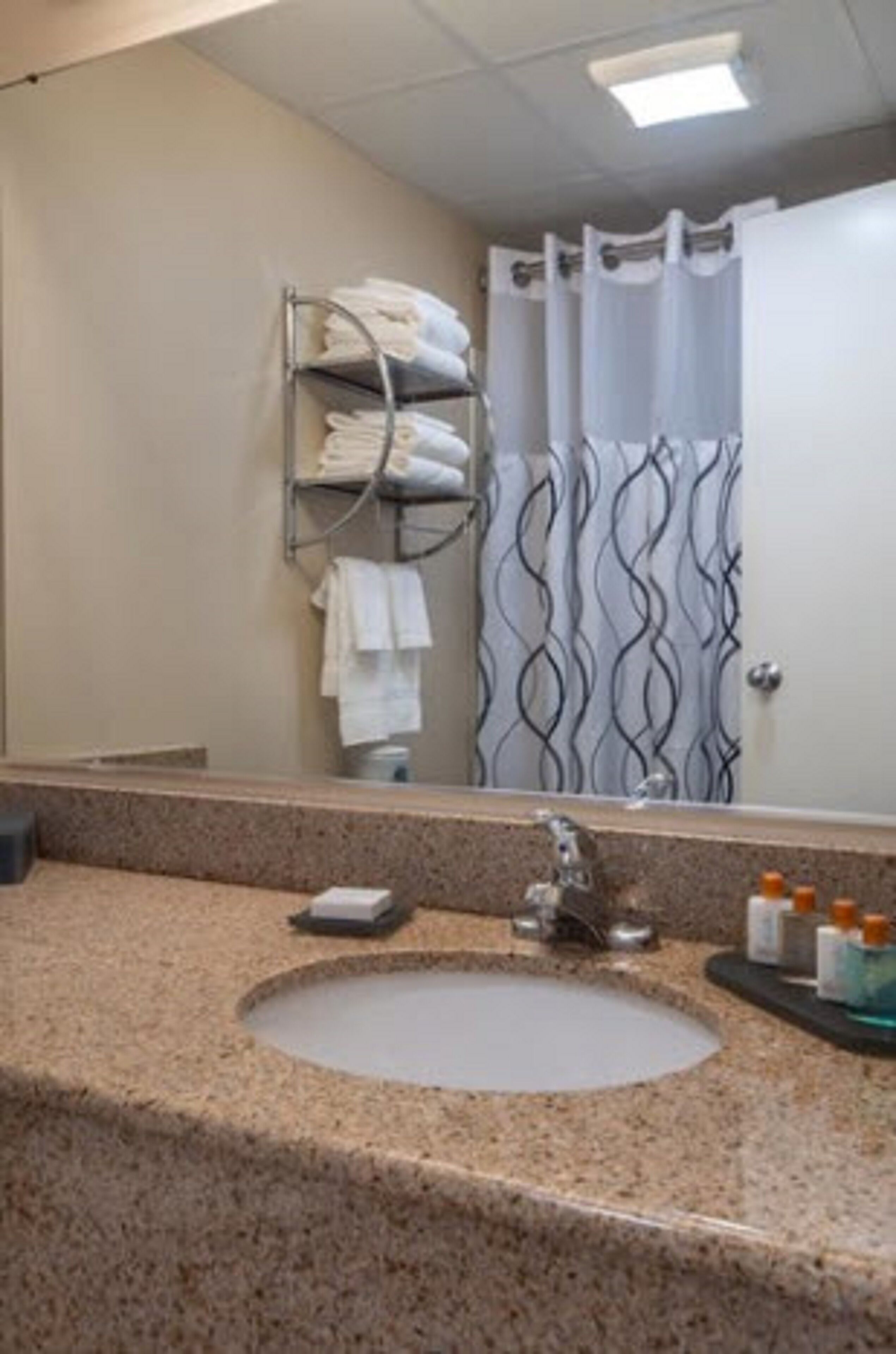 A bathroom with a granite countertop sink, a towel rack with folded towels, a shower curtain with a wavy pattern, and a mirror above the sink.