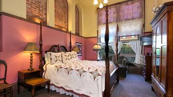 A four-poster bed with a white bedspread, a wooden nightstand with a lamp, a chair, and a dresser with a mirror.