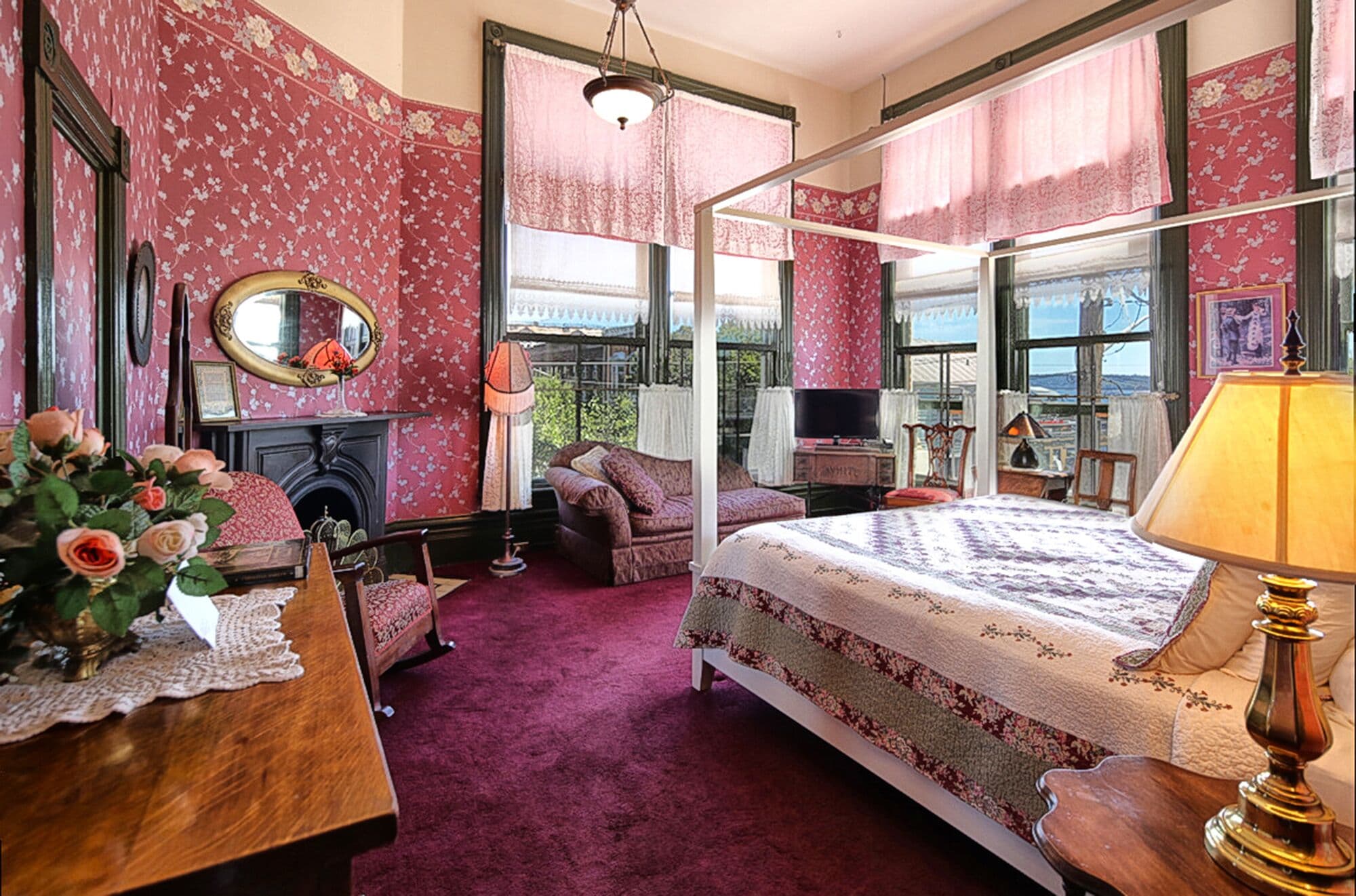 A room with a bed, a desk, a chair, a fireplace, and large windows.