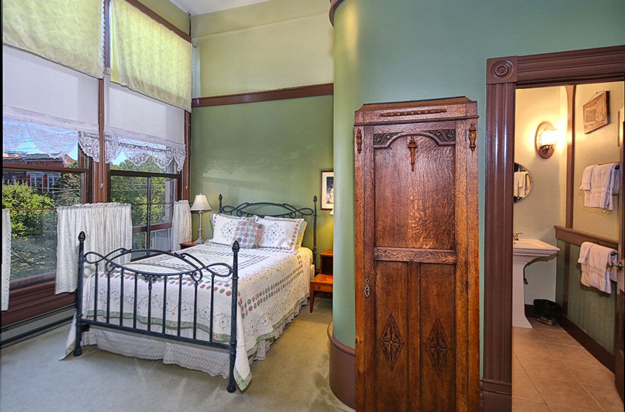 A bedroom with a bed, a wooden wardrobe, a window with lace curtains, and a view of greenery outside.